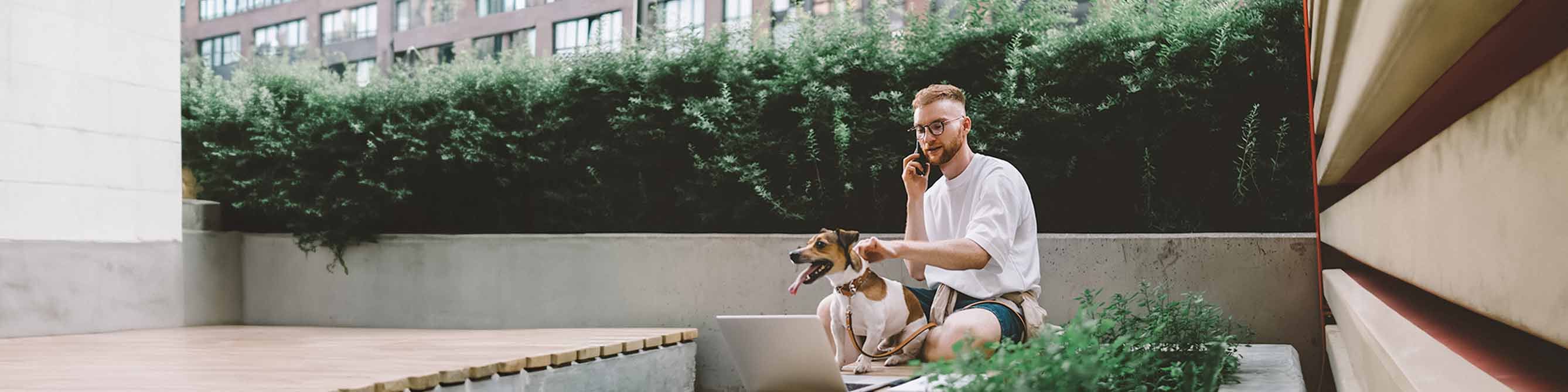 Mann sitzt draußen und telefoniert mit seinem Berater zur Hundehaftpflicht. Mit einer Hand hält er seinen Hund am Halsband fest.