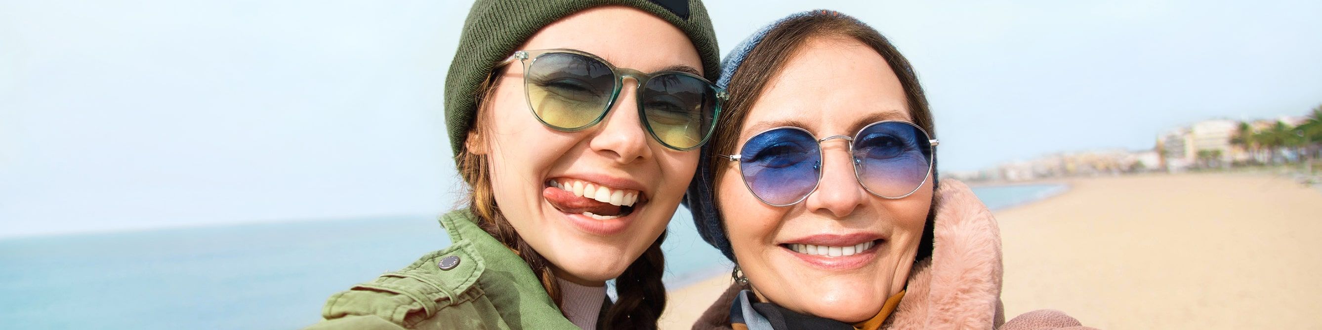Zwei Frauen am Strand machen ein Selfie und lachen in die Kamera. Beide tragen Sonnenbrillen.