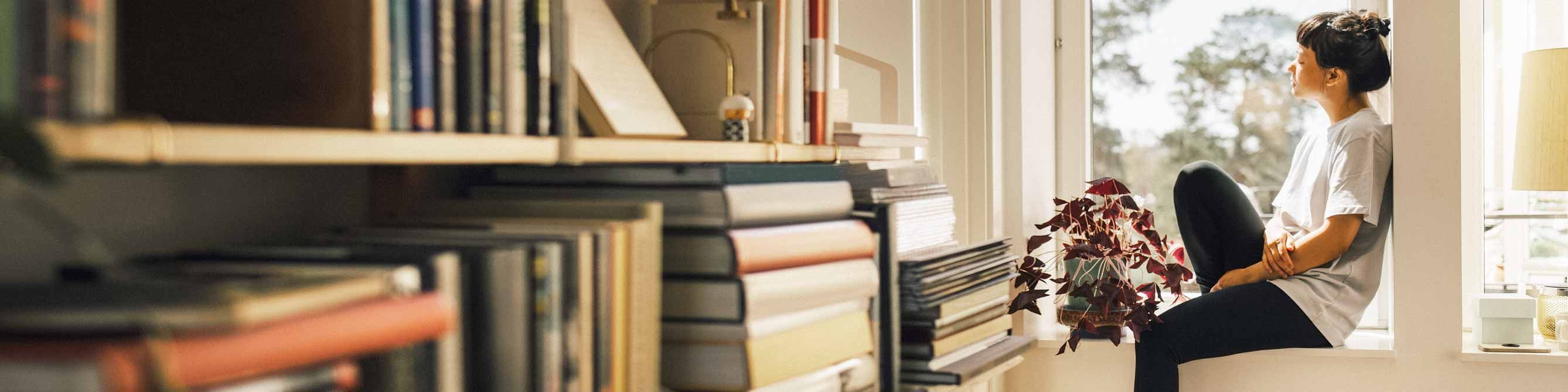 Junge Frau sitzt im Fensterrahmen und schaut hinaus, im Vordergrund sieht man ihr Bücherregal