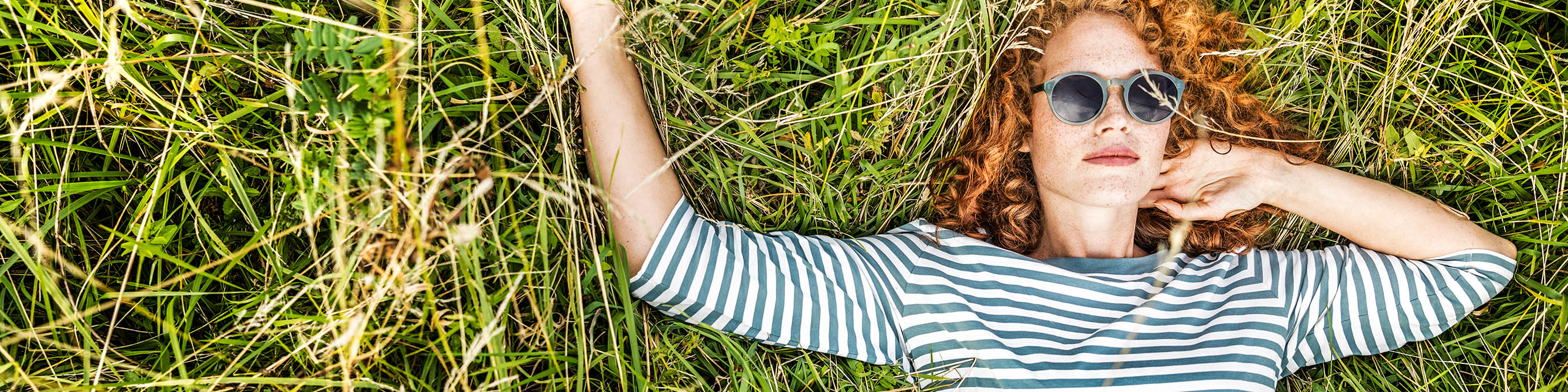 Frau mit Sonnenbrille und roten Locken liegt auf dem Rücken auf einer Wiese.