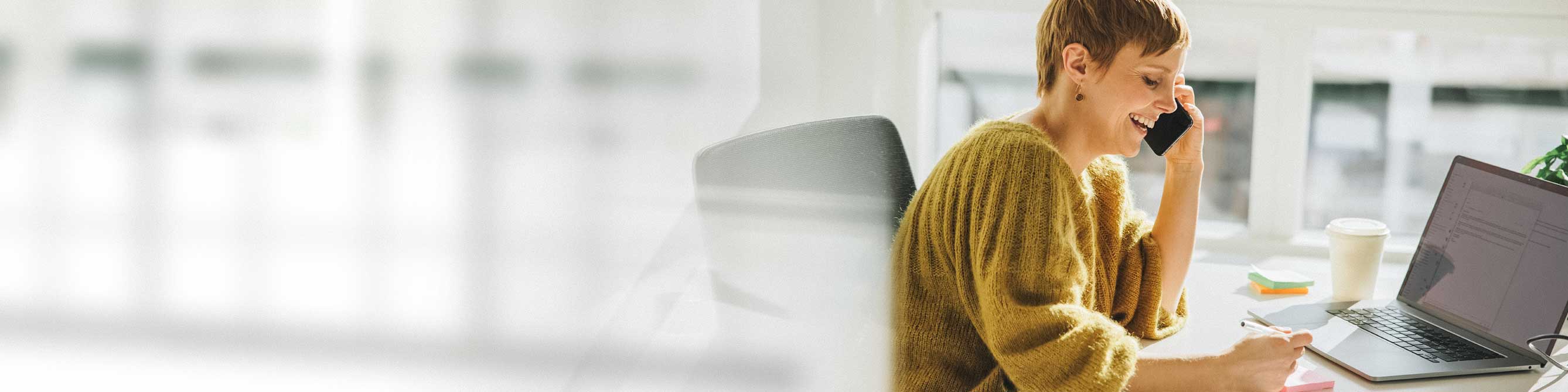 Lachende Frau im gelben Pullover mit Handy vor Laptop am Schreibtisch