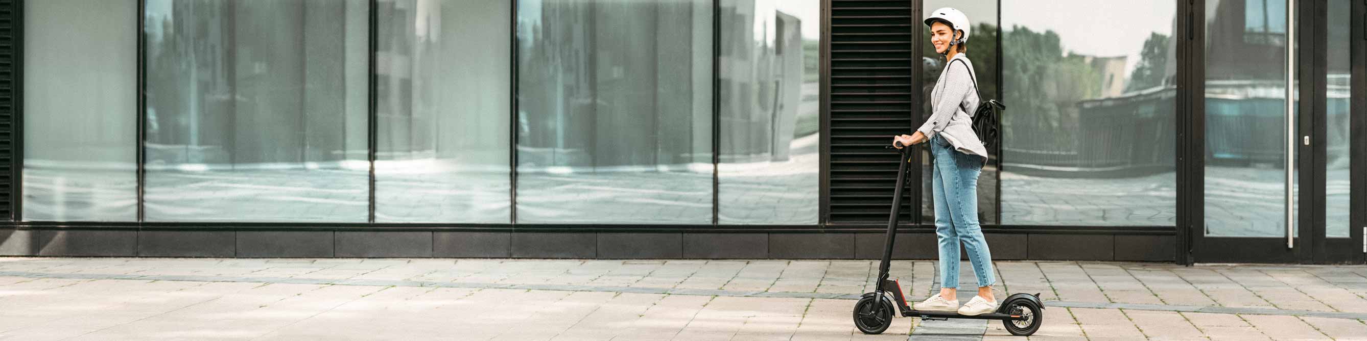 Eine blonde Frau mit Lederjacke und Schuhen mit Nieten hockt auf einem E-Scooter.