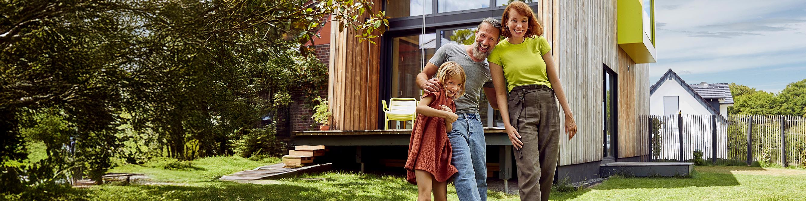 Mann, Frau und Kind stehend lachend im Garten hinter ihrem Haus und halten sich im Arm. 