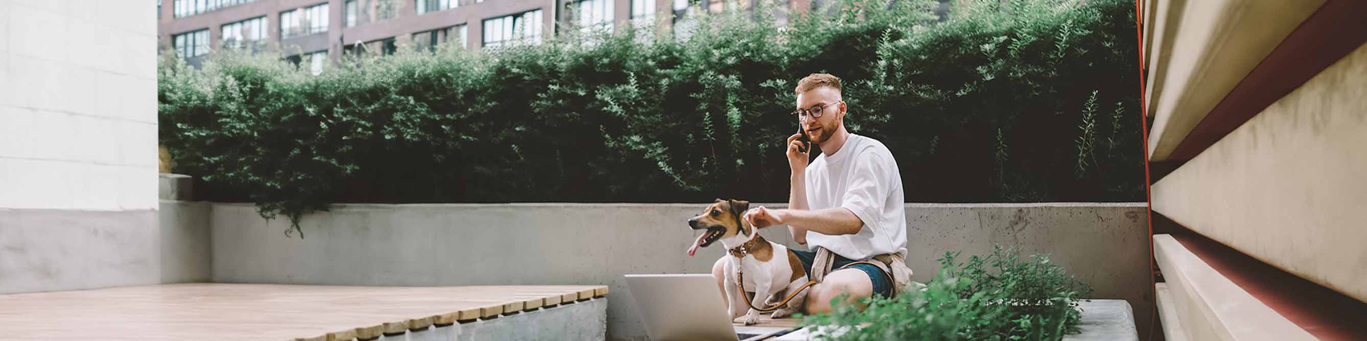 Mann sitzt draußen und telefoniert mit seinem Berater zur Hundehaftpflicht. Mit einer Hand hält er seinen Hund am Halsband fest.