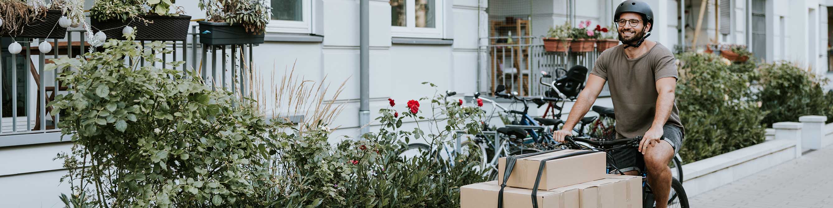 Ein Mann fährt auf einem Lastenrad, er lächelt. Auf dem Lastenrad sind vorne vier große Pakete mit Riemen gesichert. 