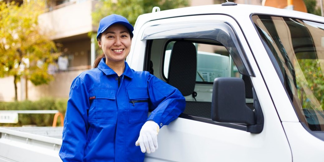 Eine Frau mit einem blauen Overall steht angelehnt an einen Lieferwagen mit Pritsche.