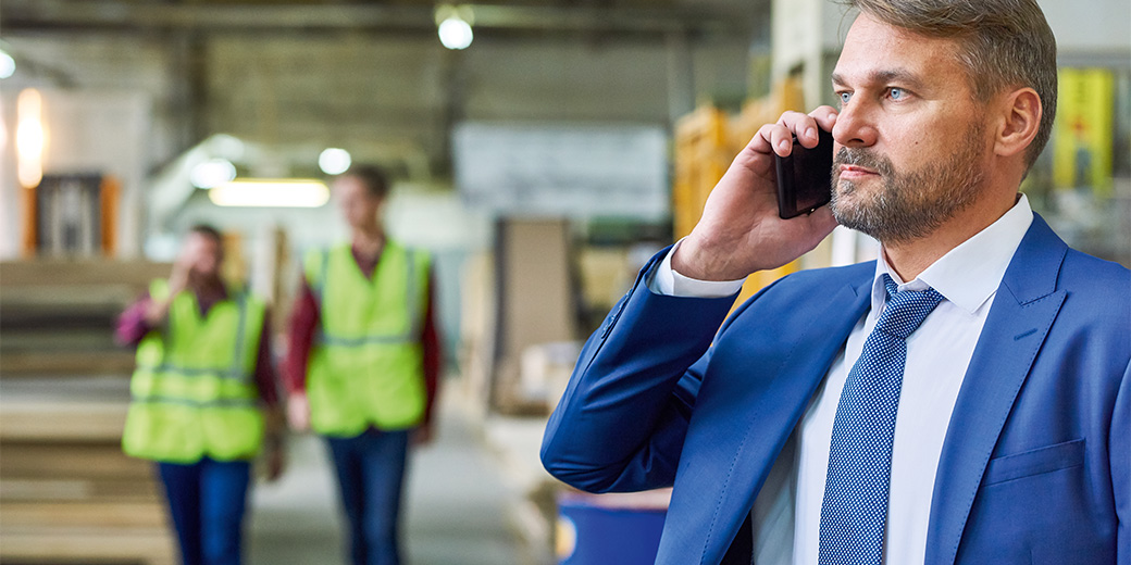 Ein Mann in Anzug und Krawatte telefoniert mit einem Handy, im Hintergrund sind zwei Personen mit Warnweste zu sehen.