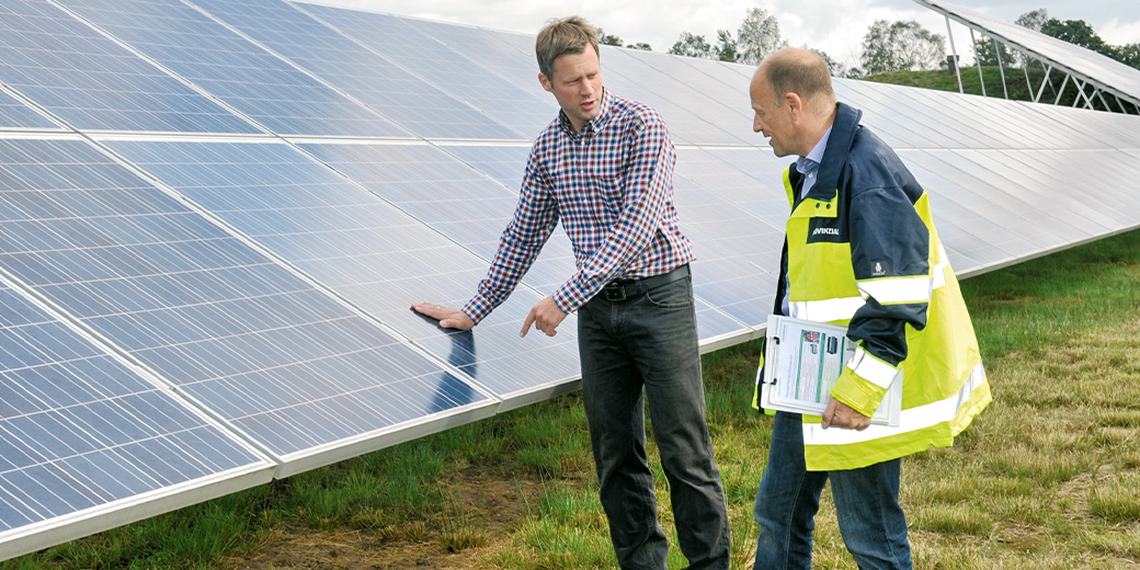 Zwei Männer, einer in einer Warnweste, stehen vor einer großen Fläche mit Solarzellen.
