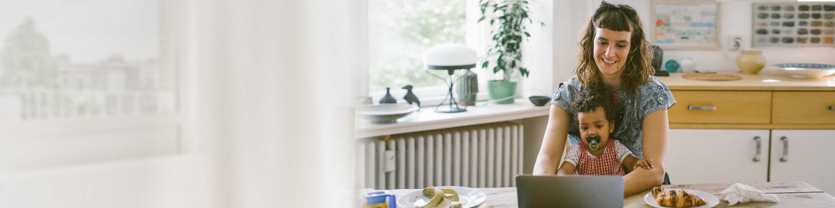 Frau sitzt am Tisch vor ihrem Laptop mit ihrem Kind auf dem Schoß