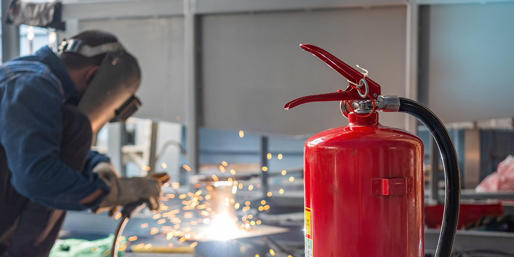 Ein Arbeiter schweißt in einer Werkshalle, im Vordergrund steht ein Feuerlöscher.