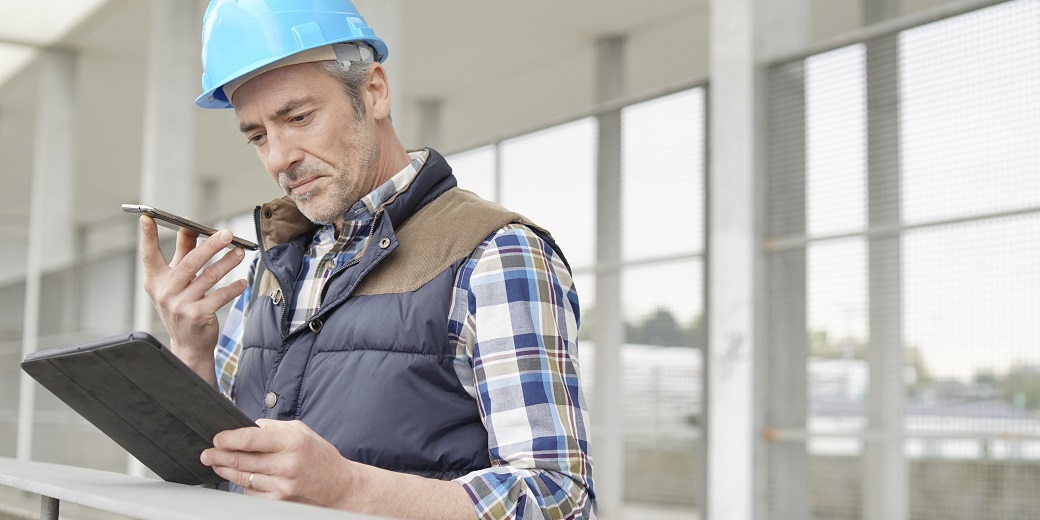 Ein Bauarbeiter mit Schutzhelm telefoniert mit seinem Handy und schaut auf ein Tablet.