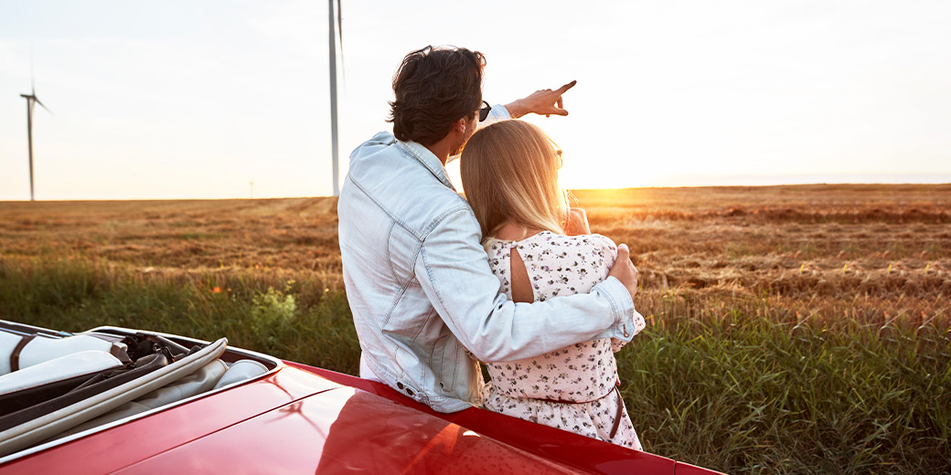 Pärchen schaut sich den Sonnenuntergang an und lehnt dabei an einem Oldtimer