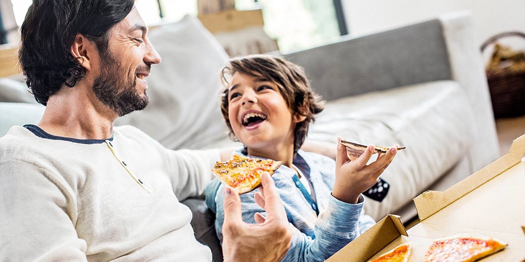 Ein Vater und sein Sohn sitzen im Wohnzimmer auf dem Sofa und essen Pizza.