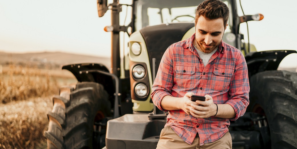 Ein Mann steht vor einem Traktor und schaut auf sein Smartphone.