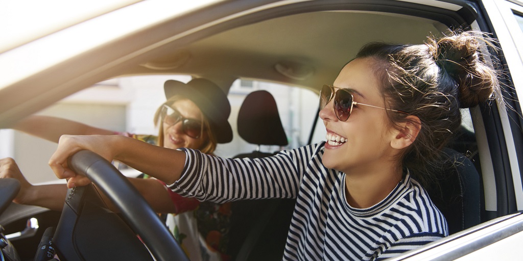 Zwei junge Frauen sitzen in einem Auto, das Seitenfenster ist geöffnet beide Frauen lachen fröhlich.