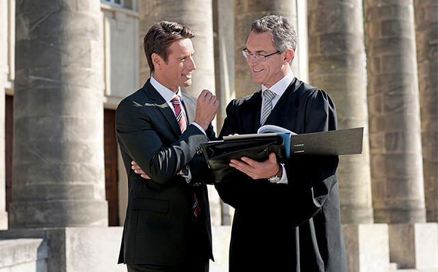 Ein Geschäftsführer bespricht sich mit seinem Anwalt; der einen Talar trägt, vor einem Gerichtsgebäude.